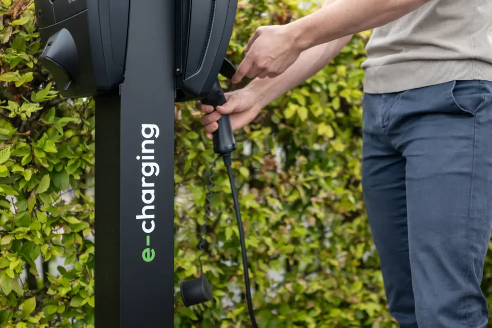 Iemand neemt de laadkabel uit een laadpaal van E-Charging bij de haag